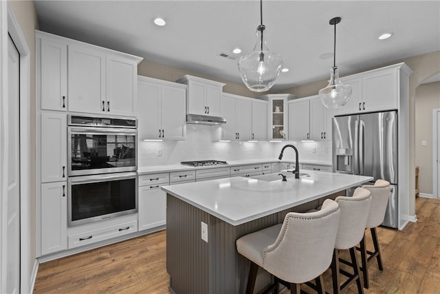 kitchen with stainless steel appliances, a kitchen island with sink, hardwood / wood-style floors, white cabinetry, and hanging light fixtures