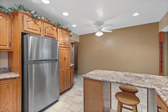 kitchen with a breakfast bar, stainless steel refrigerator, ceiling fan, and light stone counters