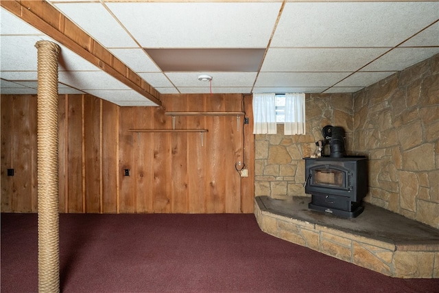 basement with carpet flooring, a wood stove, a drop ceiling, and wood walls