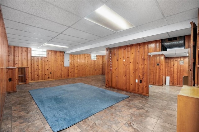 basement featuring a paneled ceiling, wood walls, and electric panel