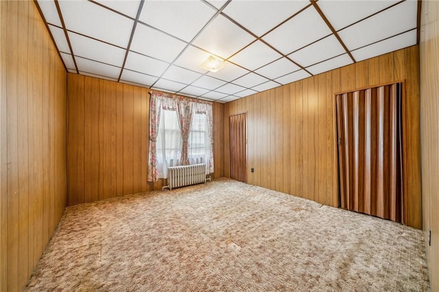 unfurnished room featuring carpet flooring, a drop ceiling, radiator, and wooden walls