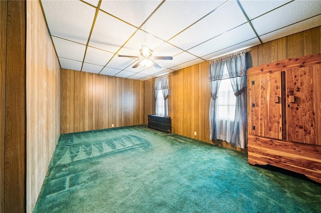 empty room with a paneled ceiling, wooden walls, ceiling fan, and dark carpet