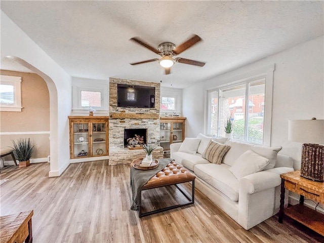 living room featuring a textured ceiling, light wood-type flooring, a stone fireplace, and ceiling fan