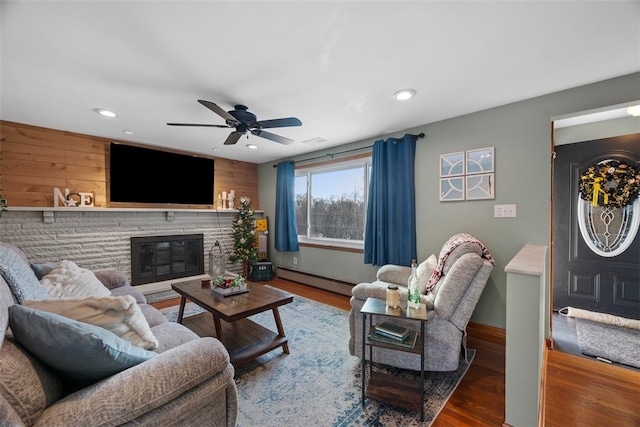 living room featuring ceiling fan, baseboard heating, a stone fireplace, wood walls, and hardwood / wood-style flooring