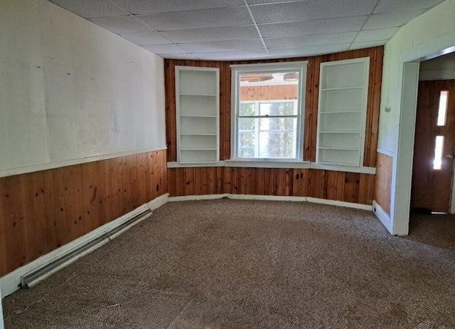 empty room featuring a paneled ceiling, carpet flooring, wood walls, and a baseboard heating unit