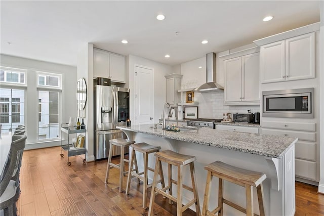 kitchen featuring white cabinetry, stainless steel appliances, wall chimney range hood, light hardwood / wood-style flooring, and an island with sink