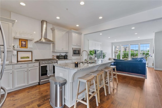 kitchen with appliances with stainless steel finishes, wall chimney range hood, white cabinets, dark hardwood / wood-style floors, and an island with sink