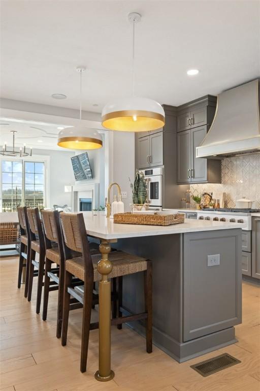 kitchen with gray cabinetry, custom range hood, an island with sink, and hanging light fixtures