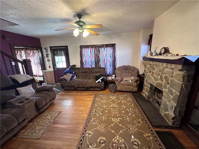 living room with wood-type flooring, a textured ceiling, a stone fireplace, and ceiling fan