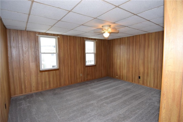 empty room featuring dark carpet, a drop ceiling, wood walls, and ceiling fan