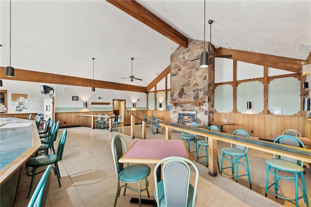 dining space featuring ceiling fan, a stone fireplace, high vaulted ceiling, beamed ceiling, and wooden walls