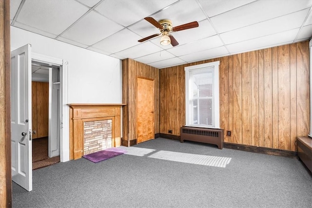 unfurnished living room featuring a paneled ceiling, dark carpet, radiator, wooden walls, and ceiling fan