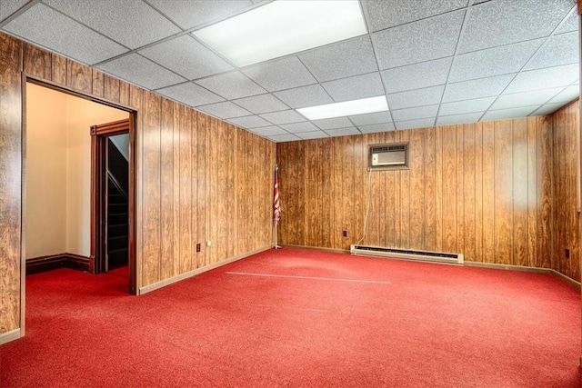 basement featuring carpet flooring, a paneled ceiling, wooden walls, and a baseboard heating unit