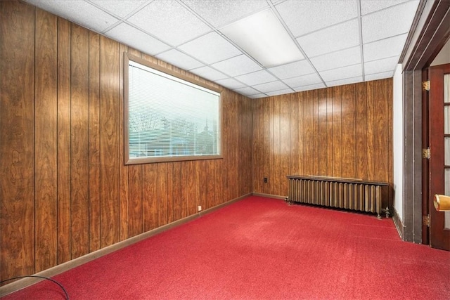 carpeted spare room featuring a drop ceiling, wood walls, and radiator heating unit