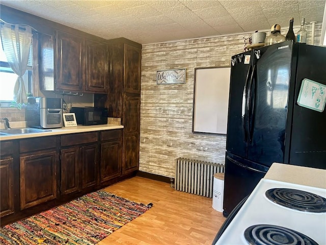 kitchen featuring dark brown cabinets, wooden walls, black appliances, radiator heating unit, and light hardwood / wood-style floors