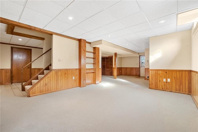 basement featuring light carpet, a paneled ceiling, and wood walls