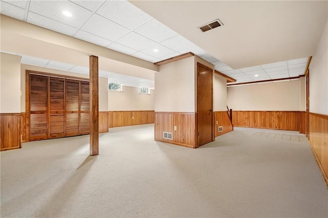 basement featuring a drop ceiling, light colored carpet, and wooden walls