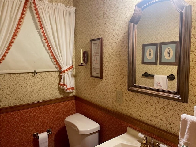 half bath with a wainscoted wall, a sink, toilet, and wallpapered walls