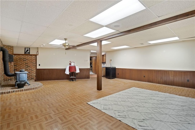 basement featuring a wood stove, ceiling fan, a drop ceiling, and light parquet floors