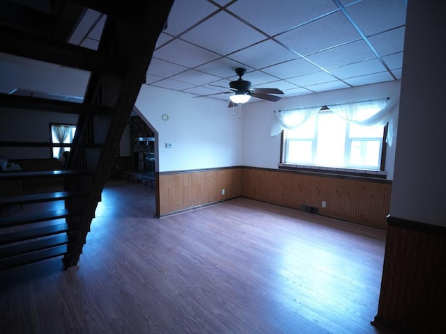 empty room with hardwood / wood-style floors, a drop ceiling, wood walls, a stone fireplace, and ceiling fan