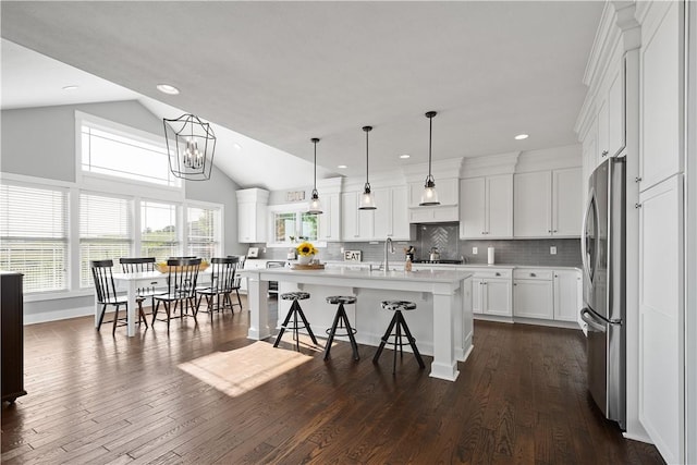 kitchen with vaulted ceiling, decorative light fixtures, white cabinetry, stainless steel refrigerator, and an island with sink