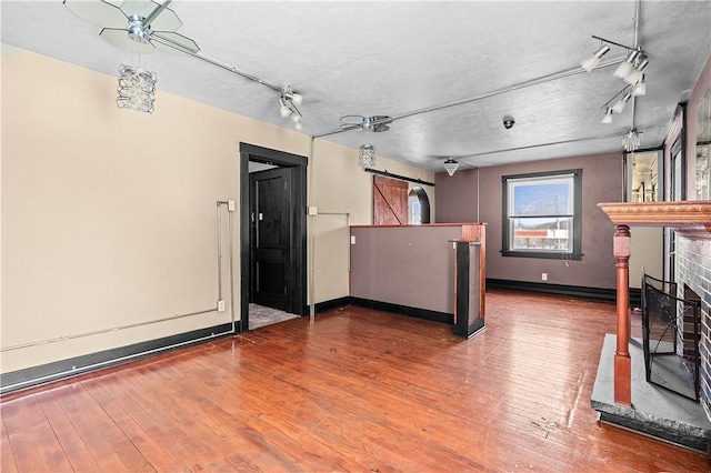 unfurnished living room featuring a barn door, wood finished floors, baseboards, a brick fireplace, and track lighting