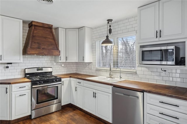 kitchen with wooden counters, sink, custom range hood, appliances with stainless steel finishes, and white cabinetry