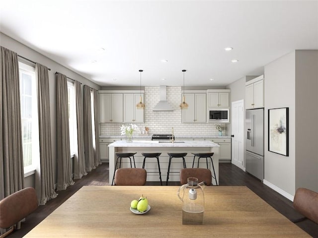 kitchen featuring stainless steel appliances, wall chimney range hood, pendant lighting, decorative backsplash, and a center island with sink