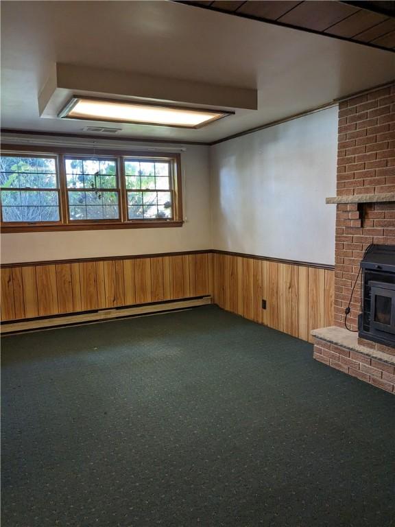 basement featuring dark colored carpet, wood walls, a wood stove, and a baseboard heating unit