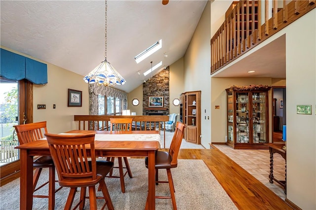 dining room with high vaulted ceiling, a stone fireplace, a skylight, light wood-type flooring, and a notable chandelier