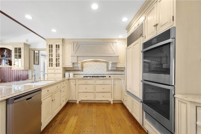 kitchen featuring sink, stainless steel appliances, premium range hood, light hardwood / wood-style floors, and cream cabinetry