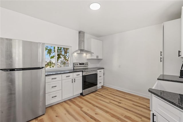 kitchen with white cabinets, wall chimney range hood, dark stone countertops, light hardwood / wood-style floors, and stainless steel appliances