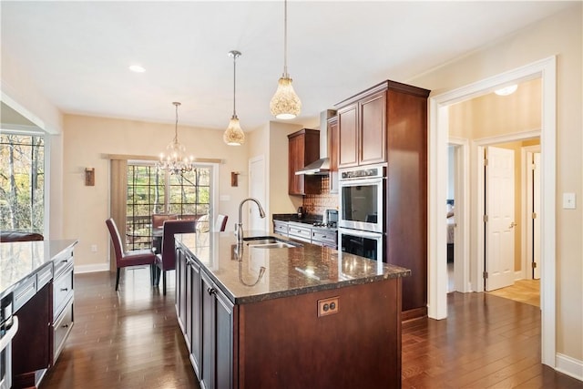 kitchen with sink, stainless steel double oven, dark stone counters, pendant lighting, and a kitchen island with sink