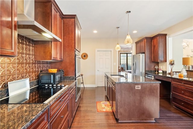 kitchen featuring sink, wall chimney exhaust hood, decorative light fixtures, a kitchen island with sink, and black appliances