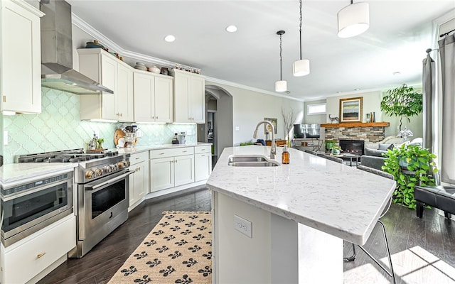 kitchen featuring light stone countertops, sink, wall chimney range hood, range with two ovens, and a kitchen island with sink