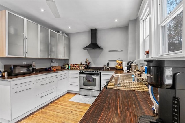 kitchen with white cabinets, wall chimney exhaust hood, light hardwood / wood-style flooring, and gas range