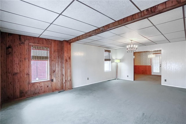 carpeted empty room with a paneled ceiling, a chandelier, and wood walls