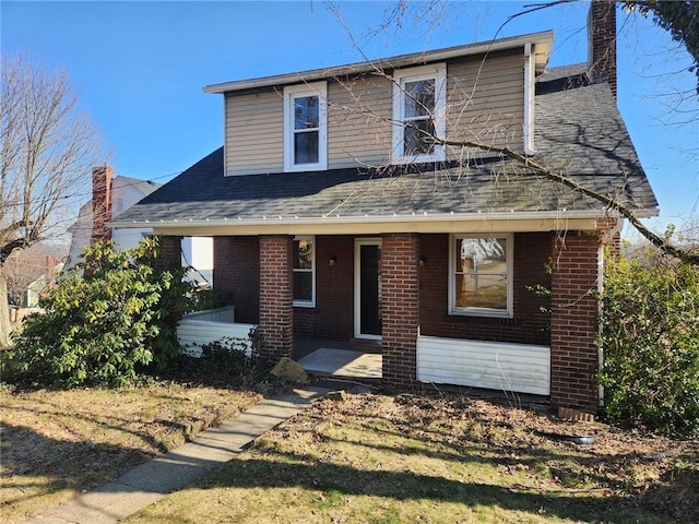 view of front facade with a chimney, brick siding, covered porch, and a shingled roof