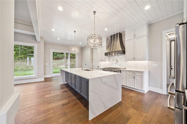 kitchen with light stone counters, custom exhaust hood, sink, white cabinetry, and an island with sink