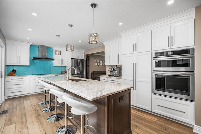 kitchen featuring stainless steel appliances, wall chimney range hood, pendant lighting, a center island with sink, and white cabinets