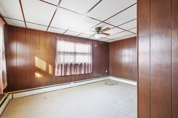 unfurnished room featuring a paneled ceiling, ceiling fan, a baseboard heating unit, and wood walls