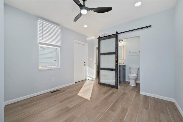 unfurnished bedroom with a barn door, ensuite bathroom, light hardwood / wood-style flooring, and ceiling fan