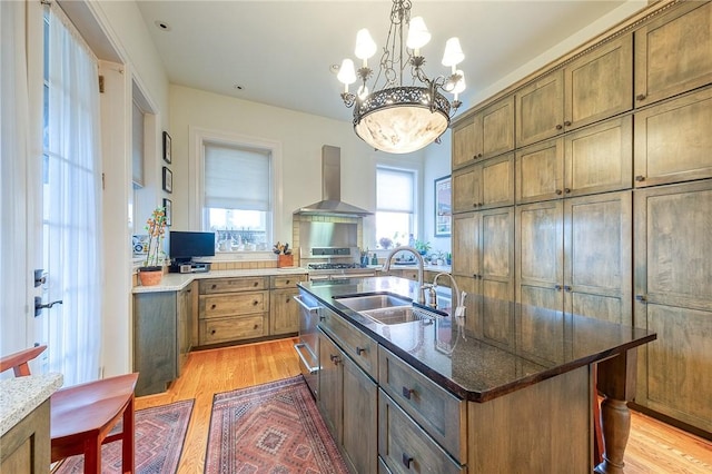 kitchen featuring pendant lighting, a kitchen island with sink, wall chimney range hood, sink, and light wood-type flooring