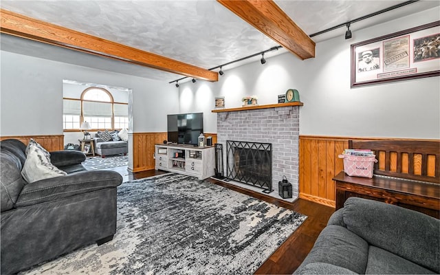 living room featuring dark wood-type flooring, rail lighting, wooden walls, a brick fireplace, and beam ceiling