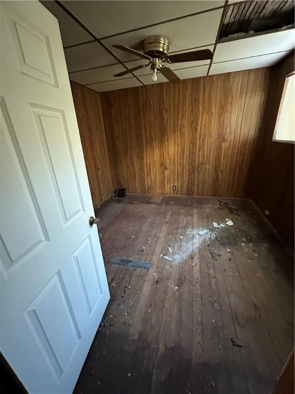 empty room with ceiling fan, wood walls, a drop ceiling, and dark hardwood / wood-style flooring