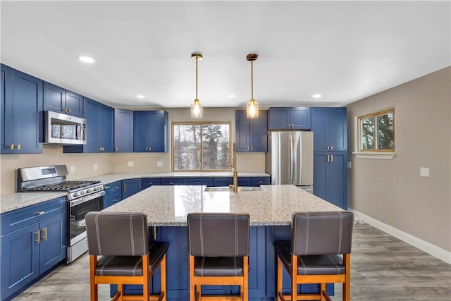 kitchen featuring appliances with stainless steel finishes, a kitchen island with sink, blue cabinets, hanging light fixtures, and light stone counters