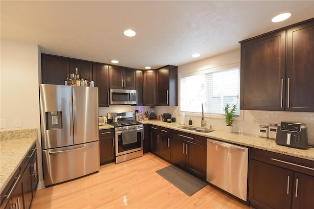 kitchen with sink, appliances with stainless steel finishes, dark brown cabinets, light stone countertops, and light hardwood / wood-style floors