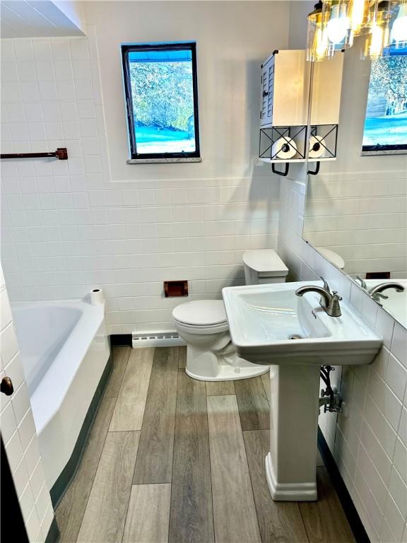 bathroom with tile walls, baseboard heating, a tub to relax in, and toilet