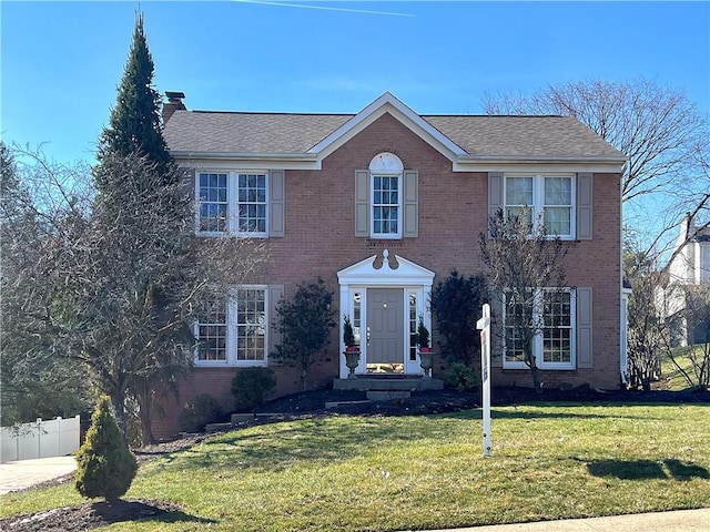 colonial inspired home with brick siding, a shingled roof, a front lawn, fence, and a chimney