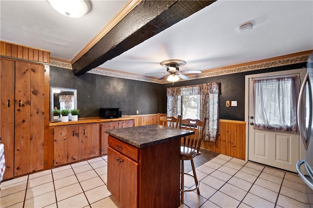 kitchen with a center island, a wainscoted wall, light tile patterned floors, dark countertops, and beamed ceiling